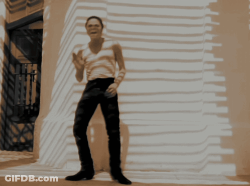 A Man In A White Tank Top And Black Pants Is Standing In Front Of A Building GIF