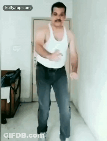 A Man In A White Tank Top And Blue Jeans Is Dancing In A Room . GIF