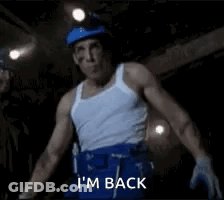 A Man In A White Tank Top And Blue Shorts Is Standing In A Dark Room And Says `` I 'M Back '' GIF