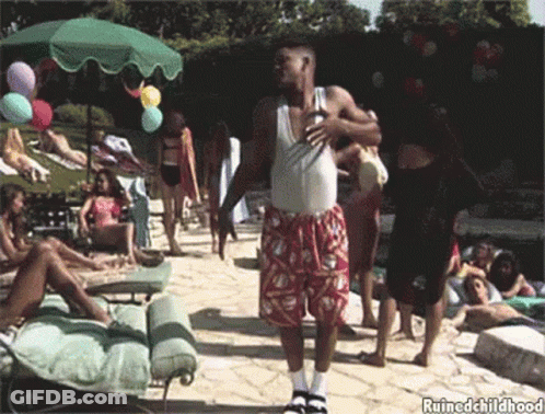 A Man In A White Tank Top And Red Shorts Is Standing In Front Of A Group Of People Near A Pool .. GIF