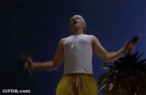 A Man In A White Tank Top And Yellow Shorts Is Standing With His Arms Outstretched In Front Of A Palm Tree . GIF