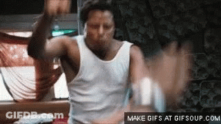 A Man In A White Tank Top Is Dancing In A Room With His Hands In The Air . GIF
