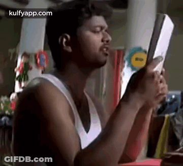 A Man In A White Tank Top Is Holding A Book In His Hands And Looking At It . GIF