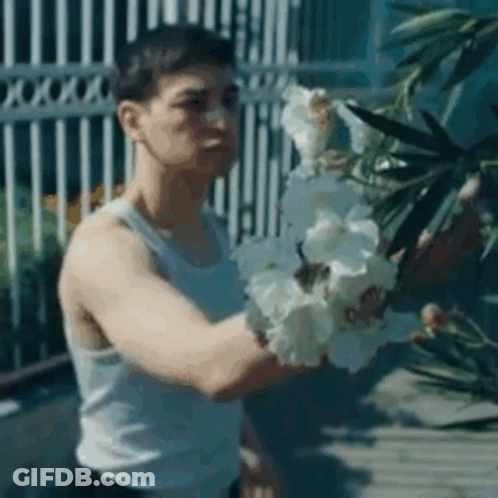 A Man In A White Tank Top Is Holding A Bunch Of White Flowers In His Hands . GIF