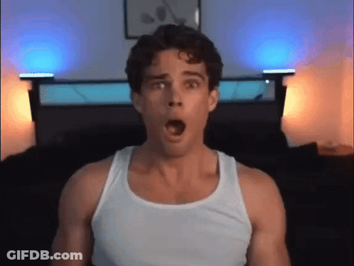 A Man In A White Tank Top Is Making A Surprised Face In Front Of A Bed GIF