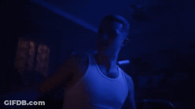 A Man In A White Tank Top Is Standing In A Dark Room With Blue Lights Behind Him . GIF