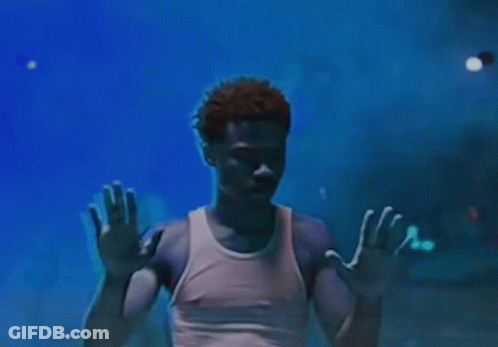 A Man In A White Tank Top Is Standing In A Dark Room With His Hands Up In The Air . GIF
