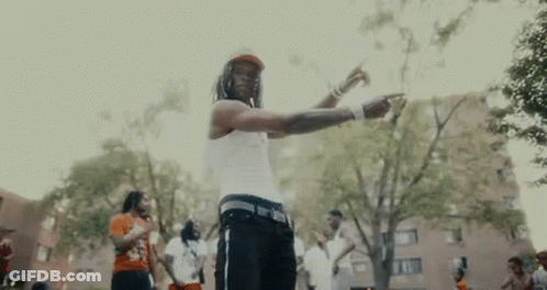 A Man In A White Tank Top Is Standing In A Park With A Group Of People . GIF