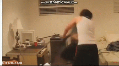 A Man In A White Tank Top Is Standing In A Room With A Computer On The Desk . GIF
