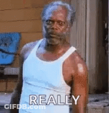 A Man In A White Tank Top Is Standing In Front Of A Building And Making A Funny Face . GIF