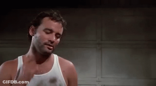 A Man In A White Tank Top Is Standing In Front Of A Garage Door And Smoking A Cigarette . GIF