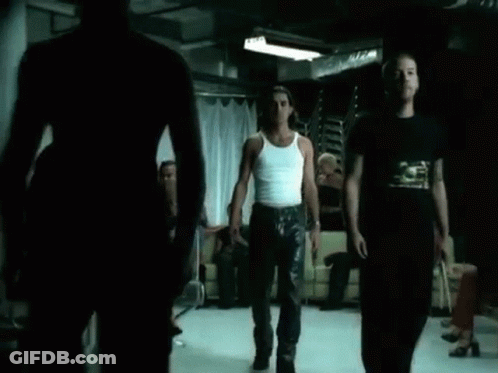 A Man In A White Tank Top Is Standing In Front Of A Group Of People In A Dark Room . GIF