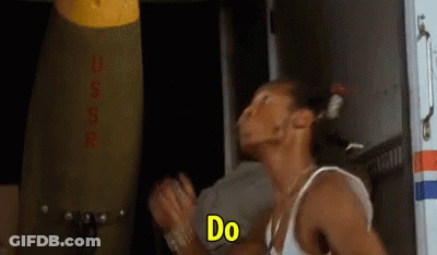 A Man In A White Tank Top Is Standing In Front Of A Large Bomb And Saying `` Do '' . GIF