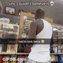 A Man In A White Tank Top Is Standing In Front Of A Store . GIF