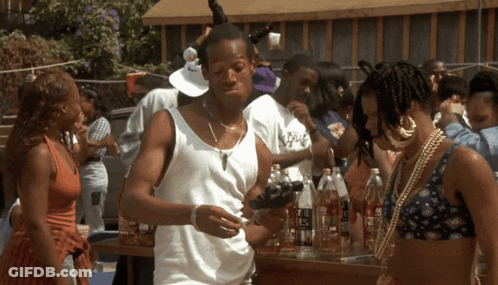 A Man In A White Tank Top Is Standing In Front Of A Table Full Of Bottles Including One That Says Es GIF