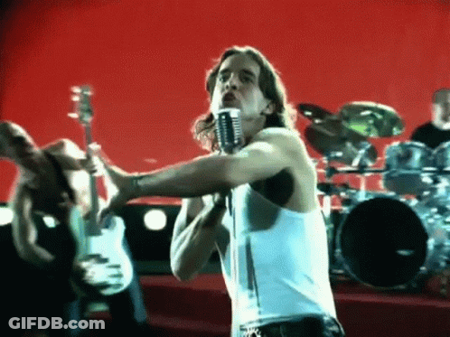 A Man In A White Tank Top Singing Into A Microphone In Front Of A Drum Set . GIF