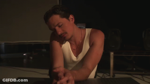 A Man In A White Tank Top Sits At A Desk GIF