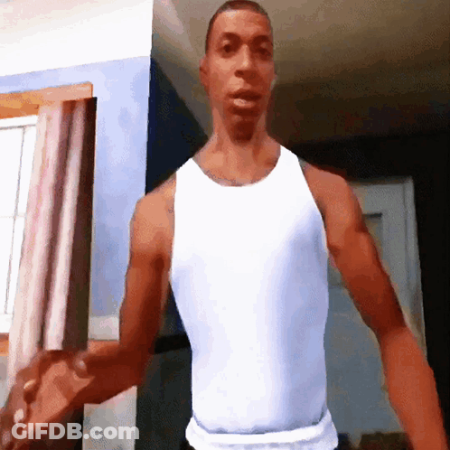 A Man In A White Tank Top Stands In A Room GIF