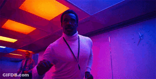 A Man In A White Turtleneck Is Dancing In A Pink Room With A Lanyard Around His Neck . GIF
