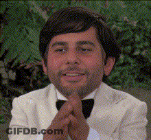 A Man In A White Tuxedo And Bow Tie Is Smiling With His Hands Folded GIF