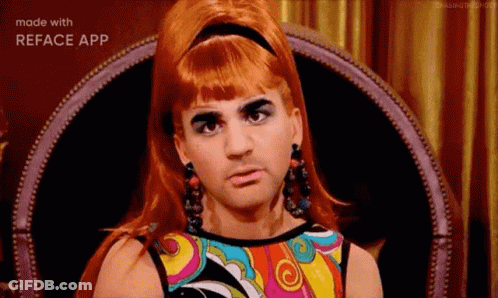 A Man In A Wig And Earrings Is Sitting In A Chair With A Colorful Dress On . GIF