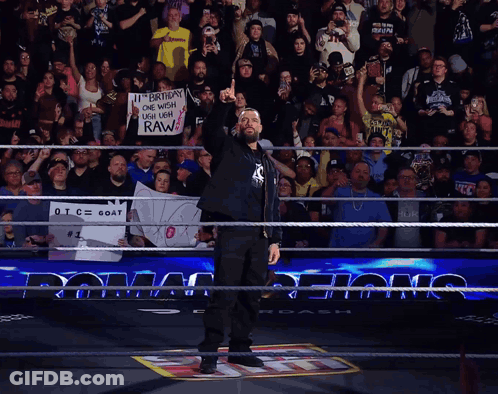 A Man In A Wrestling Ring Holds Up His Fist In Front Of A Crowd Holding A Sign That Says Raw GIF