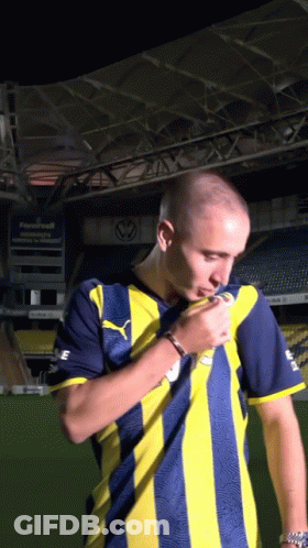 A Man In A Yellow And Blue Striped Shirt Stands In Front Of A Stadium GIF