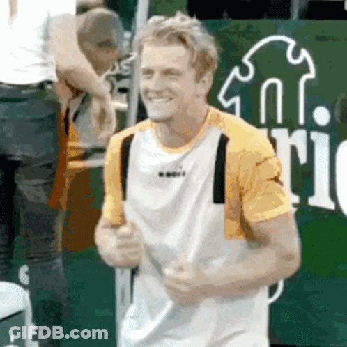 A Man In A Yellow And White Shirt Is Smiling While Holding A Tennis Racket . GIF