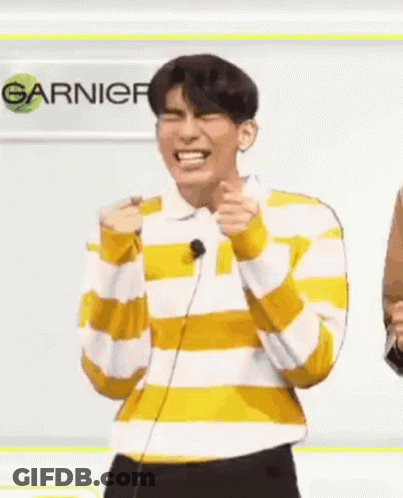 A Man In A Yellow And White Striped Shirt Is Laughing While Standing In Front Of A Microphone And A Sign . GIF