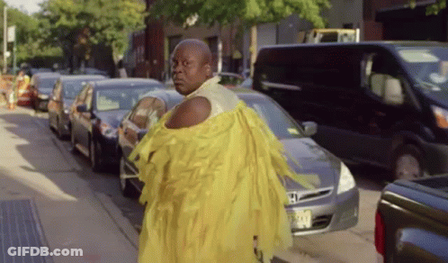 A Man In A Yellow Dress Is Walking Down A Busy Street GIF
