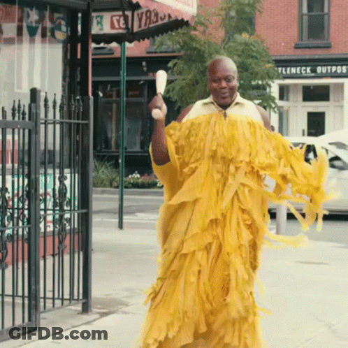 A Man In A Yellow Dress Is Walking Down The Street In Front Of A Store Called Ttleneck Outpost GIF