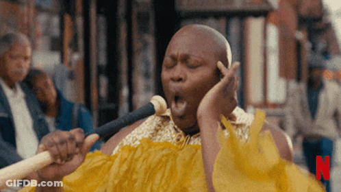 A Man In A Yellow Dress Singing Into A Microphone With The Letter N In The Corner GIF
