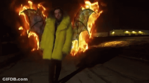 A Man In A Yellow Fur Coat Is Standing In Front Of Flames . GIF