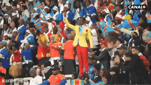 A Man In A Yellow Jacket And Red Pants Stands In Front Of A Crowd With Canal + Written On The Bottom Right Corner GIF
