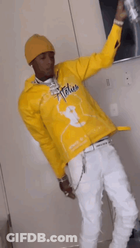 A Man In A Yellow Jacket And White Pants Is Standing In A Room Holding A Can Of Soda . GIF