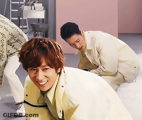 A Man In A Yellow Jacket Is Smiling While Another Man Squats Behind Him GIF
