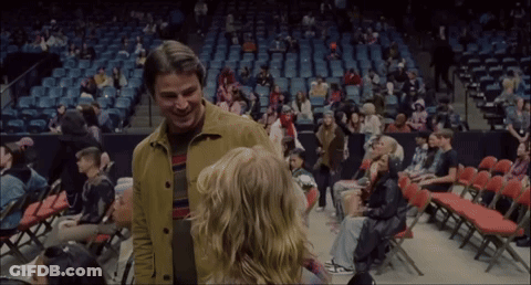 A Man In A Yellow Jacket Stands Next To A Woman In A Stadium GIF