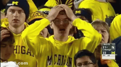 A Man In A Yellow Michigan Sweatshirt Holds His Head In His Hands GIF