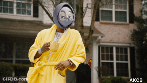 A Man In A Yellow Robe Is Holding A Cup Of Coffee In Front Of A House GIF