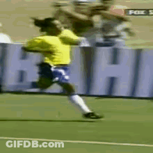 A Man In A Yellow Shirt And Blue Shorts Is Running On A Soccer Field In Front Of A Sign That Says Fox GIF