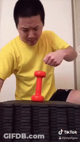 A Man In A Yellow Shirt Holds A Red Dumbbell On A Tire GIF