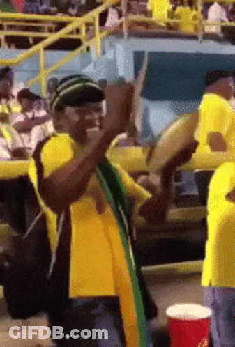 A Man In A Yellow Shirt Is Playing A Drum In A Crowd GIF