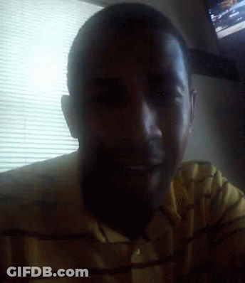 A Man In A Yellow Shirt Is Smiling In Front Of A Window With Blinds GIF