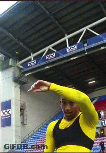 A Man In A Yellow Shirt Is Standing In Front Of A Stadium With A Sign That Says Vv On It GIF