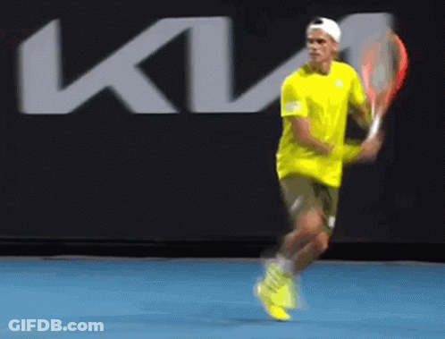 A Man In A Yellow Shirt Is Swinging A Tennis Racket In Front Of A Kia Logo GIF
