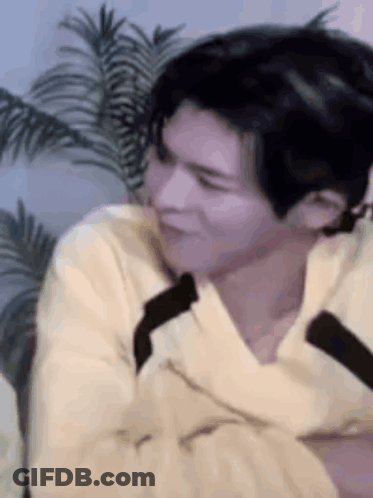 A Man In A Yellow Sweater Is Sitting In Front Of A Palm Tree And Making A Funny Face . GIF