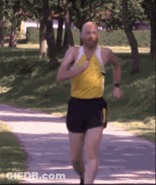 A Man In A Yellow Tank Top And Black Shorts Is Running Down A Path In A Park GIF