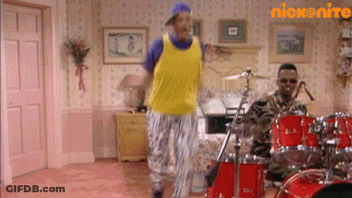 A Man In A Yellow Tank Top Is Dancing In Front Of A Drum Set In A Room . GIF