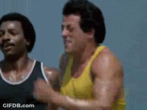 A Man In A Yellow Tank Top Is Running Next To Another Man . GIF
