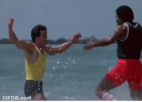 A Man In A Yellow Tank Top Is Standing Next To Another Man In Red Shorts On The Beach GIF